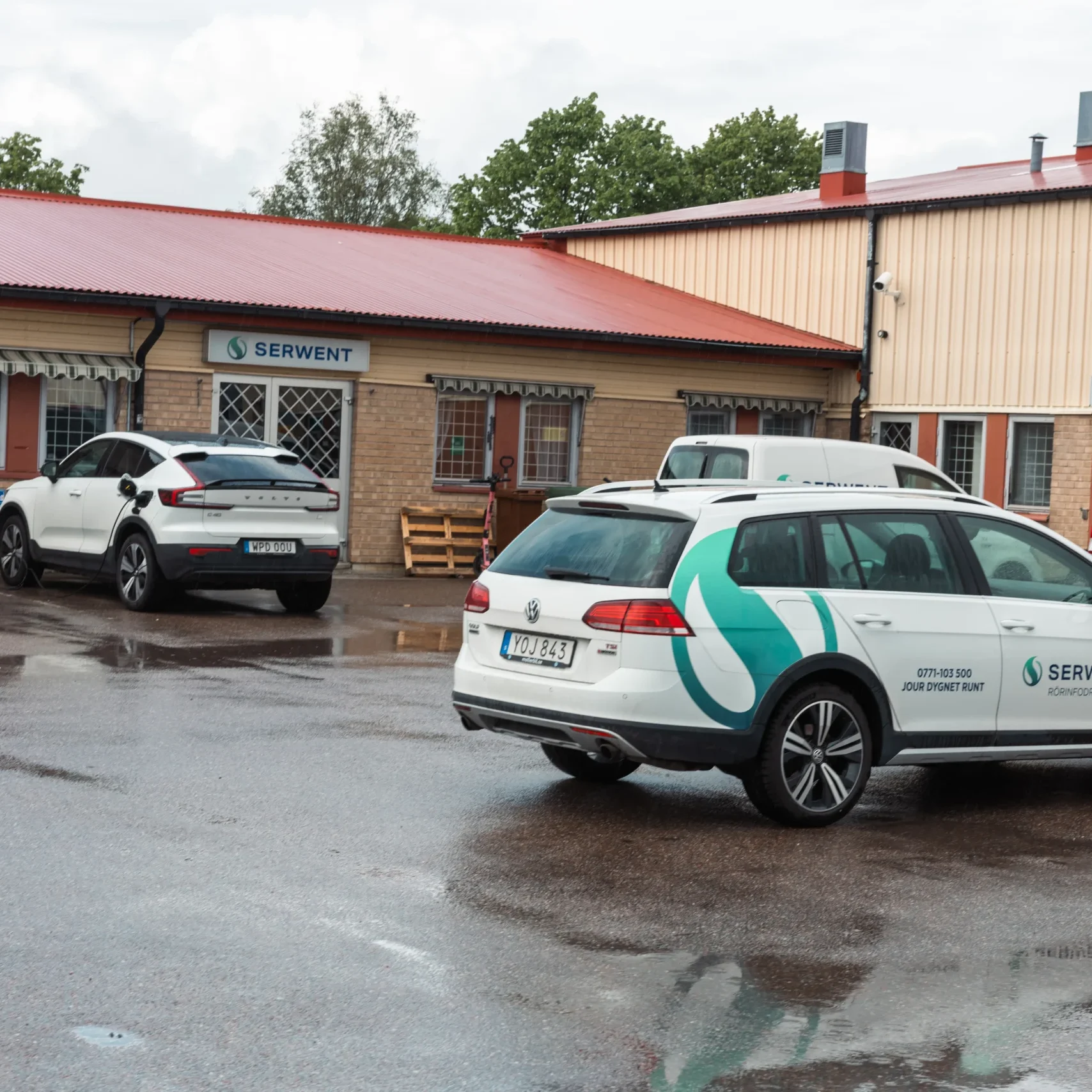 Serwent Västerås Tre bilar, varav två elbilar med Serwient-märkning, står parkerade utanför en byggnad med rött tak och Serwient- och Förråd-skyltar i Serwent Västerås. Den våta marken tyder på att det nyligen har regnat.