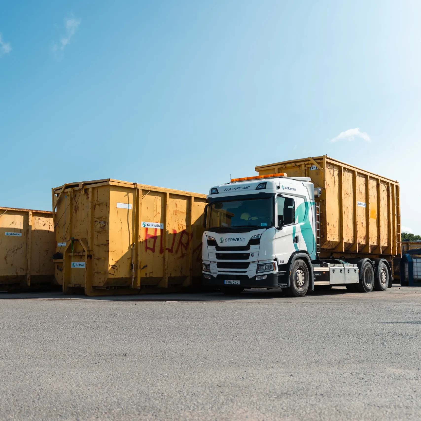 Fordon Uppsala En vit Serwent Uppsala-semitrailer med grön logotyp står parkerad bredvid stora gula industricontainrar på en asfalterad tomt under en klarblå himmel.