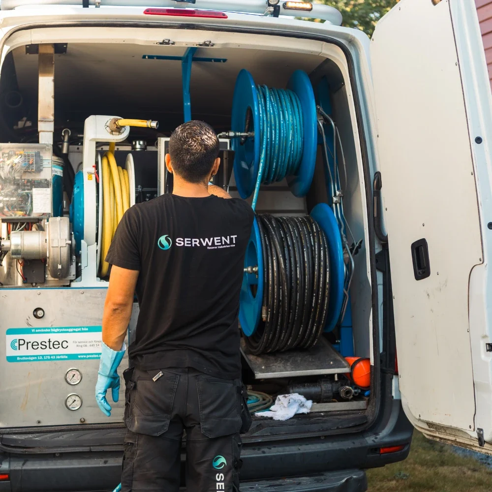 En man i svart Serwent-skjorta står vid den öppna baksidan av en servicebil och hanterar blå och svarta slangar och utrustning inne i fordonet.