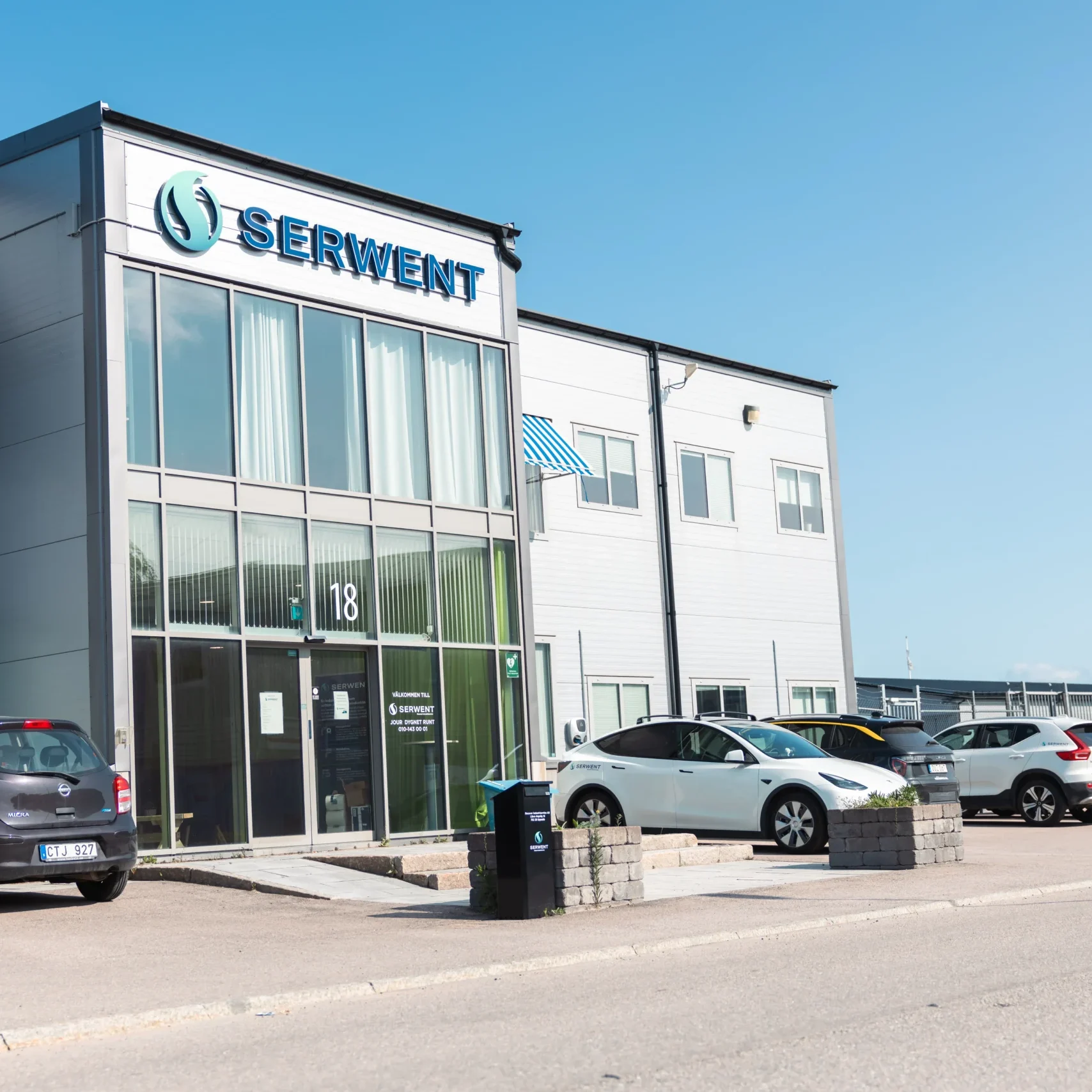 Uppsala En modern kontorsbyggnad i två våningar med stora fönster och en skylt med texten "SERWENT" ovanför entrén står i solskenet. Flera bilar står parkerade utanför och hälsar besökarna välkomna till Serwent Stockholm.