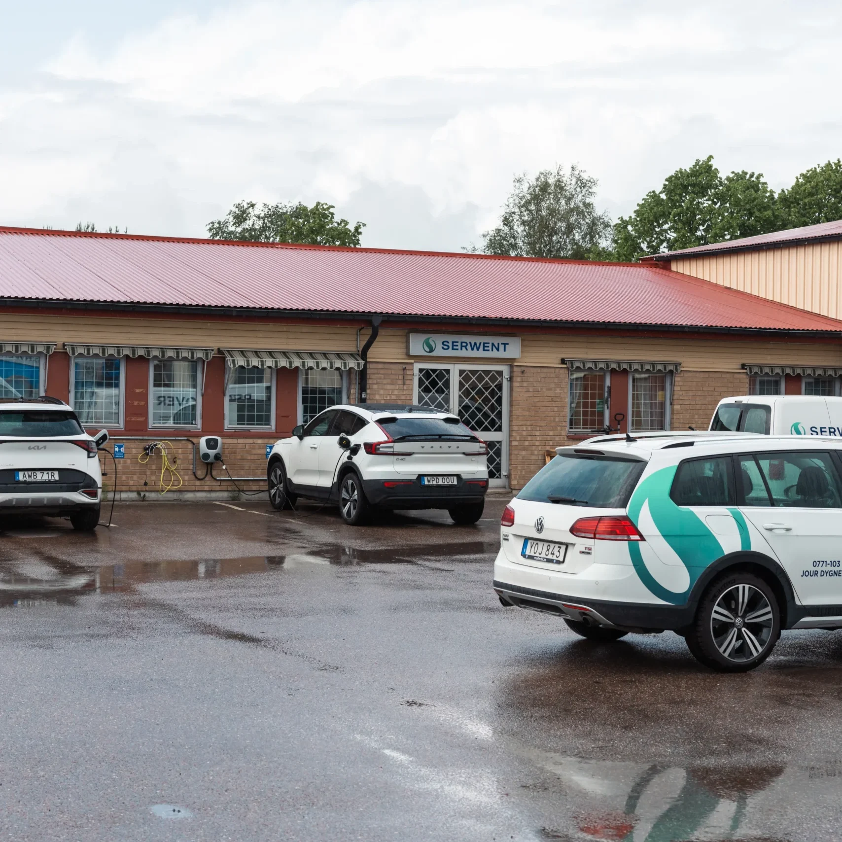 Serwent Västerås En liten kontorsbyggnad med rött tak syns på bilden en regnig dag i Serwent Västerås. Flera vita bilar, vissa med företagslogotyper, står parkerade utanför. Den våta marken reflekterar bilarna och byggnaden.