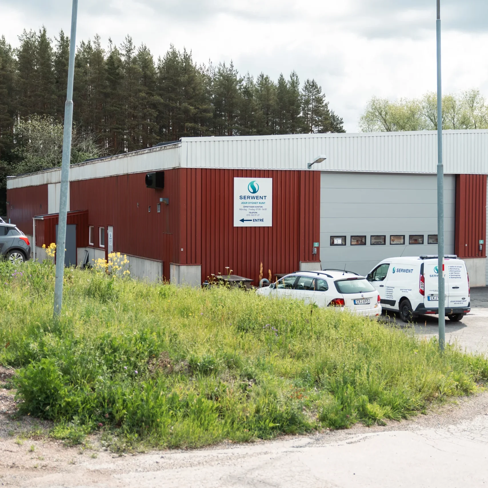 Serwent Gävle En röd industribyggnad med vitt tak och flera garageportar, där Serwent Uppsala har sitt säte, är omgiven av bilar och skåpbilar. Vilda blommor fyller den gräsbevuxna förgrunden, och i bakgrunden syns en skog under en molnig himmel.