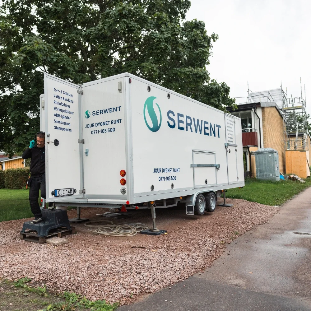 relining4 En vit servicetrailer från Serwent står parkerad på en grusväg nära en byggnad och bredvid den står en man i svarta arbetskläder. Träd och byggnadsställningar syns i bakgrunden - perfekt för projekt som kräver relining. Vad är relining?.