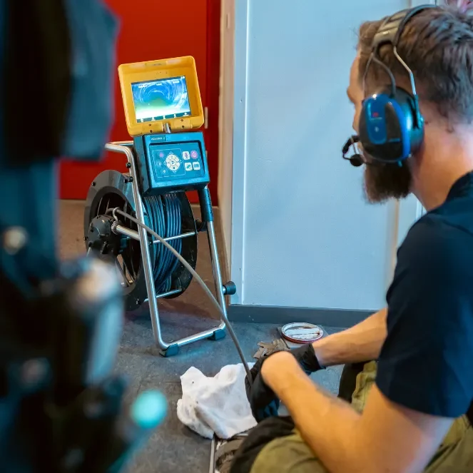 rorinspektion.brf En man med hörlurar och handskar sitter bredvid en monitor som är ansluten till en kabelrulle och inspekterar ett videoflöde från en kamera, troligen för inspektion av rör eller avlopp, med en röd vägg i bakgrunden.