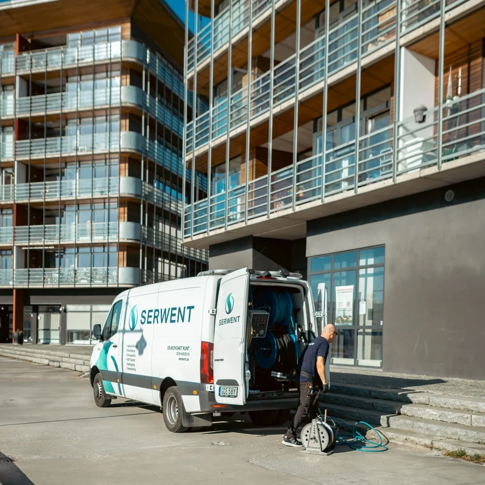stamspolning En arbetare står vid en vit Serwent-bil med städutrustning, parkerad utanför ett modernt hyreshus med balkonger och stora fönster en solig dag.