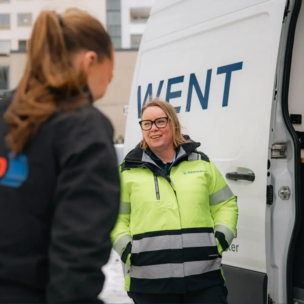 uppdrag serwent isab Två personer står utomhus bredvid en vit Serwent-skåpbil. Den ena, som bär glasögon och en gul säkerhetsväst, ler medan hen pratar med den andra, som är klädd i mörka kläder, med byggnader synliga i bakgrunden.