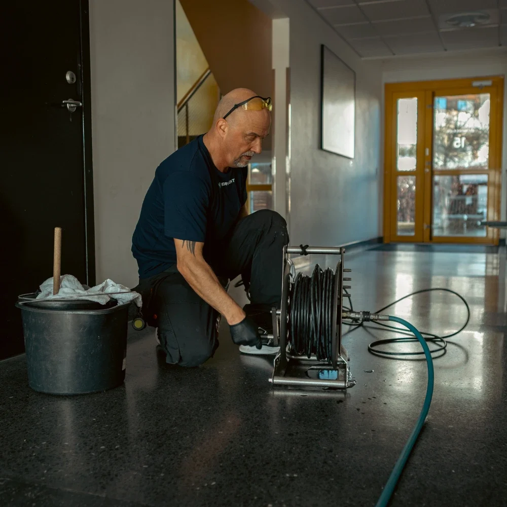 En man står på knä på golvet i en korridor och förbereder en slangvinda med svarta slangar för stamspolning. Han bär handskar och skyddsglasögon på huvudet. En närliggande hink innehåller rengöringsmedel, medan gula dörrar lyser upp den väl upplysta korridoren. Så går stamspolning till.